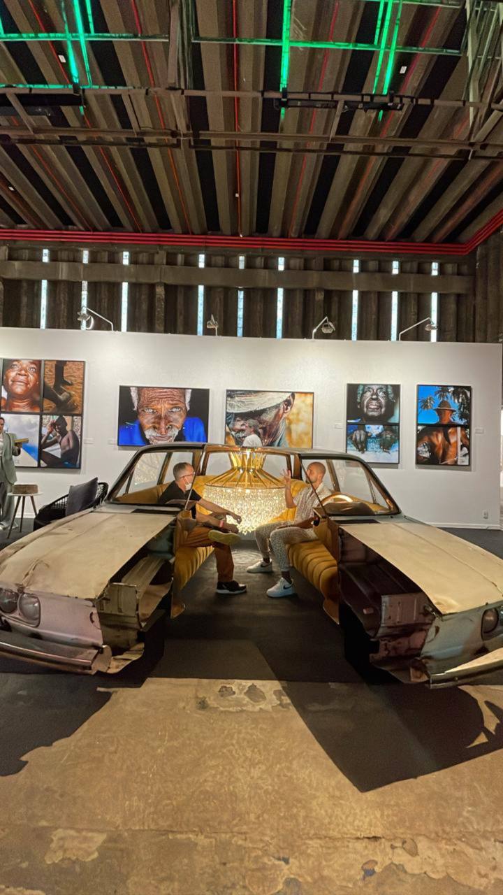 Instalação artística com carros e pessoas dentro, obras de arte na parede ao fundo